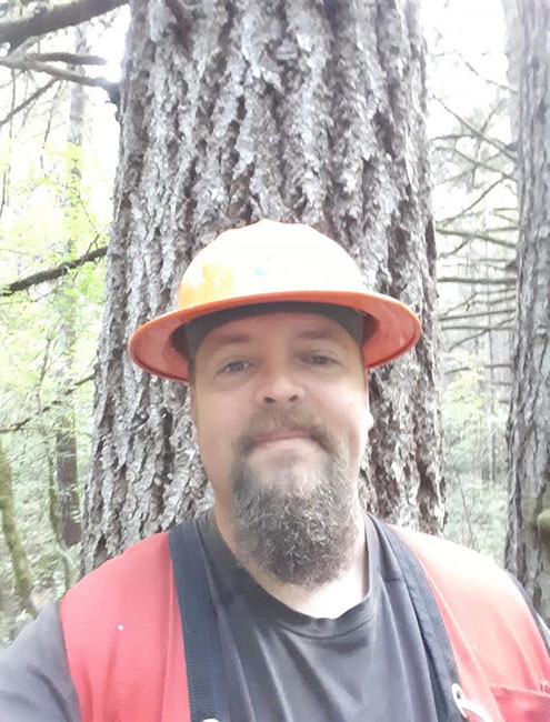 Eric DeWitt looking serious, wearing an orange hard hat standing in front of a tree.