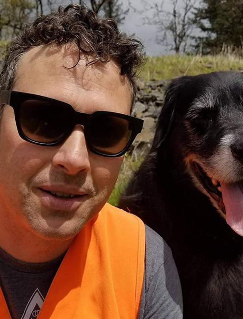 Thomas Dee selfie outside in sunglasses and an orange vest next to black dog with white snout.