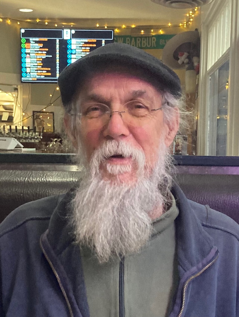 Wade Bullier white bearded, with cap and glassees smiling in restaurant.