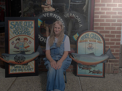 White woman in jean overalls sitting on the sidewalk between two painted salmon signs of her creation.