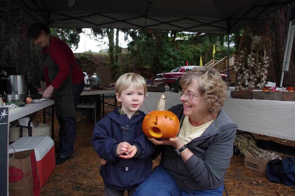 A white blonde haired boy looks at camera while a white older woman shows him a small jack'o'lantern.